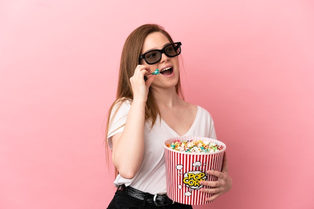 Teenager-Mädchen über isoliertem rosa Hintergrund mit 3D-Brille und hält einen großen Eimer Popcorn