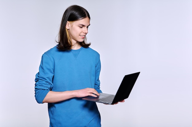 Teenager-Männlicher Student mit Laptop, der gegen einen leichten Studio-Hintergrund posiert, hübscher Typ mit modischer Frisur mit Computer in den Händen, Jugend-Technologie-Lebensstil, Konzept für junge Menschen