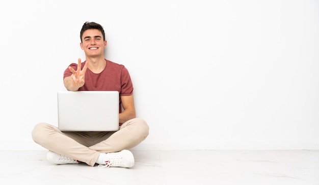 Teenager Mann sitzt auf dem Boden mit seinem Laptop glücklich und zählt drei mit den Fingern