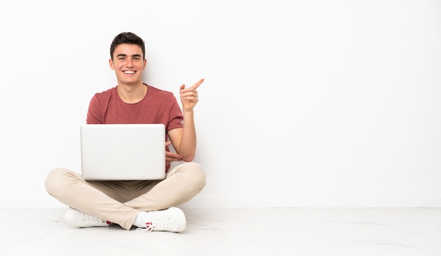 Teenager Mann sitzt auf der Flor mit seinem Laptop zeigt Finger zur Seite