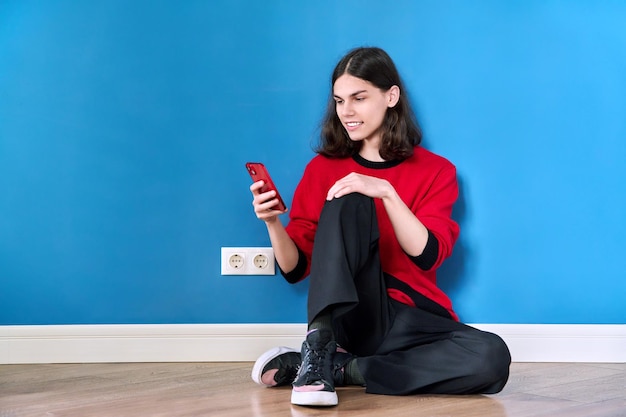 Teenager mit Smartphone auf blauem Hintergrund auf dem Boden sitzend