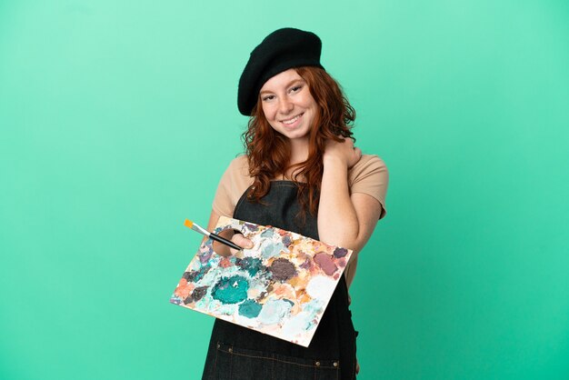 Teenager rothaarige Künstler hält eine Palette isoliert auf grünem Hintergrund lachen