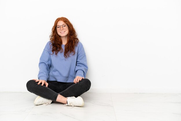 Foto teenager rothaarige mädchen auf dem boden sitzend auf weißem hintergrund mit brille und lächelnd