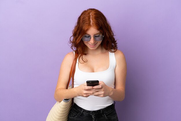 Teenager rothaarige Mädchen hält eine Strandtasche isoliert auf lila Hintergrund Senden einer Nachricht mit dem Handy