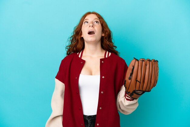 Teenager rothaarige Mädchen mit Baseballhandschuh isoliert auf blauem Hintergrund nach oben schauend und mit überraschtem Ausdruck