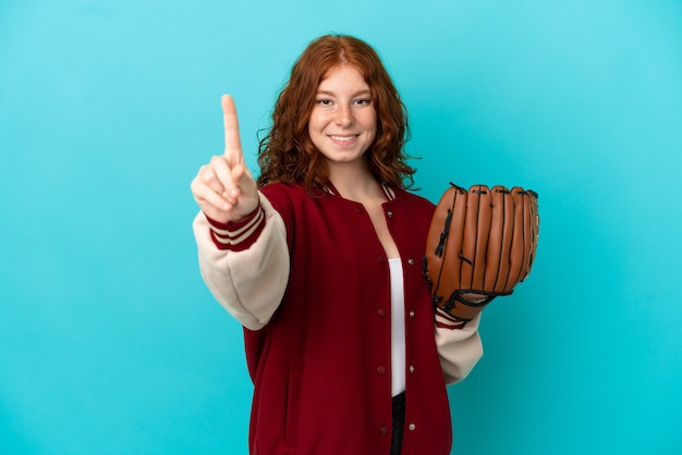 Teenager rothaarige mädchen mit baseballhandschuh isoliert auf blauem hintergrund zeigt und hebt einen finger