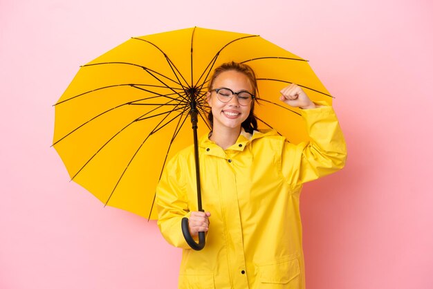 Teenager-Russin mit regenfestem Mantel und Regenschirm isoliert auf pinkfarbenem Hintergrund mit starker Geste