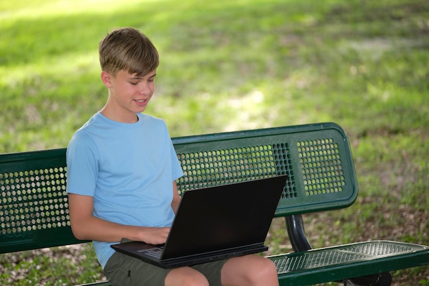 Teenager-Schuljunge, der auf einem Laptop-Computer tippt, der an sonnigen Sommertagen auf einer Bank im Freien sitzt Bildung während des Quarantänekonzepts