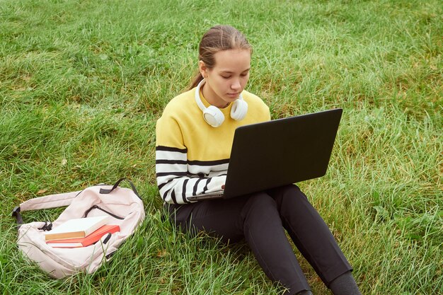 Teenager-Student benutzt einen Laptop, während er auf dem Gras sitzt und sich auf Prüfungen vorbereitet