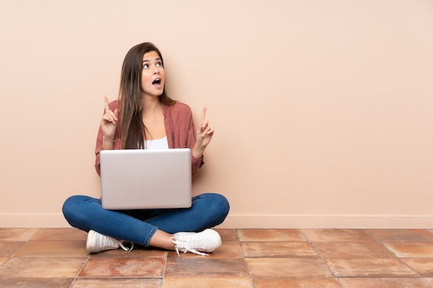 Teenager-Student sitzt auf dem Boden mit einem Laptop überrascht und zeigt nach oben