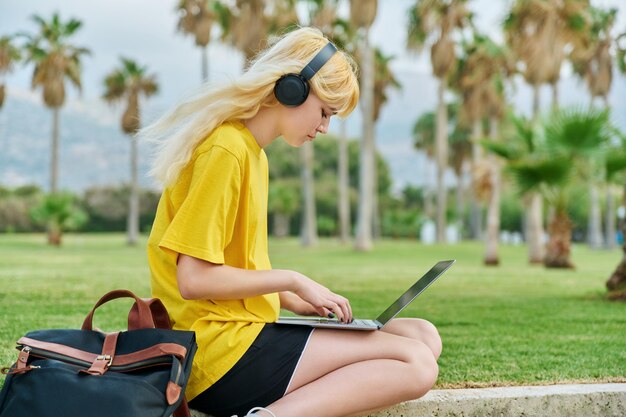 Teenager-Studentin mit Kopfhörern, die mit Laptop im Park sitzt