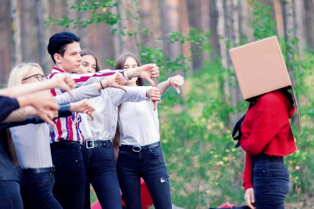 Teenager zeigen eine Fingergeste auf das Mädchen mit einer Kiste auf dem Kopf Gruppe von Teenagern im Wald