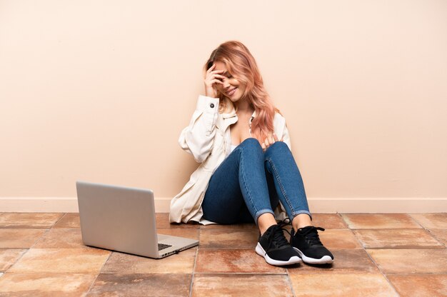 Teenagerfrau mit einem Laptop, der auf dem Boden beim Innenlachen sitzt