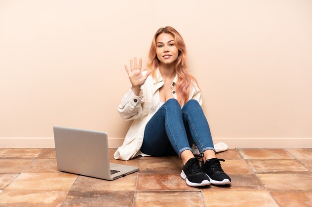 Teenagerfrau mit einem Laptop, der auf dem Boden drinnen sitzt und fünf mit den Fingern zählt