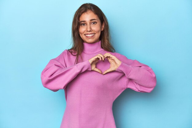 Teenagerin in gemütlichem rosa Sweatshirt auf blau lächelnd und mit Händen eine Herzform zeigend