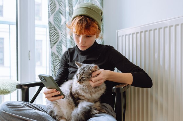 Teenagerin sitzt auf einem Stuhl, hält eine Katze und schaut auf das Telefon