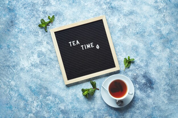 Teezeit schwarzes Buchstabenbrett mit Text auf blauem Tisch mit Glastasse Tee mit Minzblättern