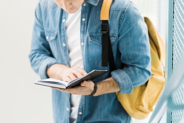 Teilansicht einer Studentin im Jeanshemd mit gelbem Rucksack, die ein Notizbuch liest