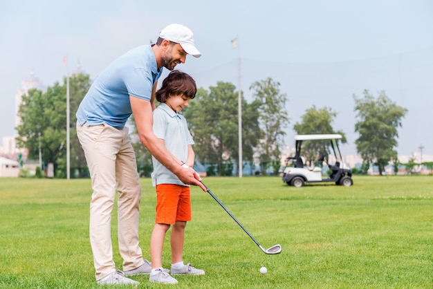 Teilen mit Golferfahrung. Fröhlicher junger Mann, der seinem Sohn das Golfspielen beibringt, während er auf dem Golfplatz steht