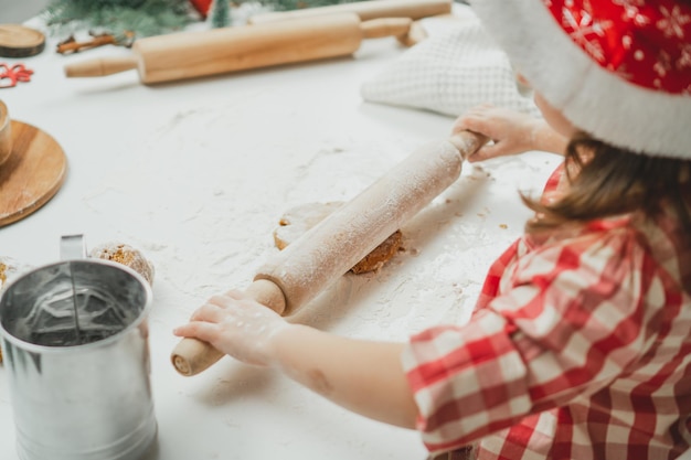 Teilweise verschwommene Hände eines kleinen Mädchens in Weihnachtsmütze und kariertem Hemd rollen Teig für Kekse mit Nudelholz aus
