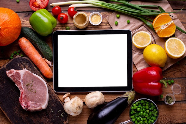 Tela vazia do tablet eletrônico branco temperado com carne e vegetais diferentes em um fundo de madeira