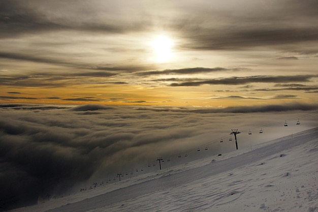 Foto teleférico com cadeiras acima das nuvens durante o pôr do sol, a escuridão se aprofunda abaixo