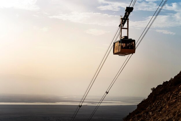 Teleférico con gente bajando en funicular sobre montañas en el desierto de Judea en el fondo del Mar Muerto Vista panorámica del ferrocarril por cable con el cielo brumoso y el mar en el horizonte Israel Masada