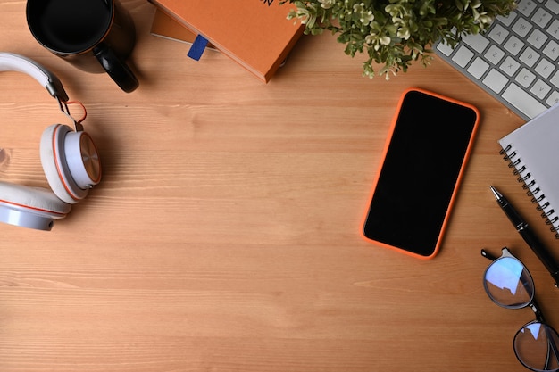 Telefone celular, fone de ouvido, caderno, óculos, xícara de café e planta de casa em fundo de madeira.