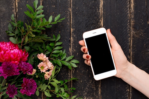 Telefone celular na mão feminina e um buquê de flores em uma madeira preta