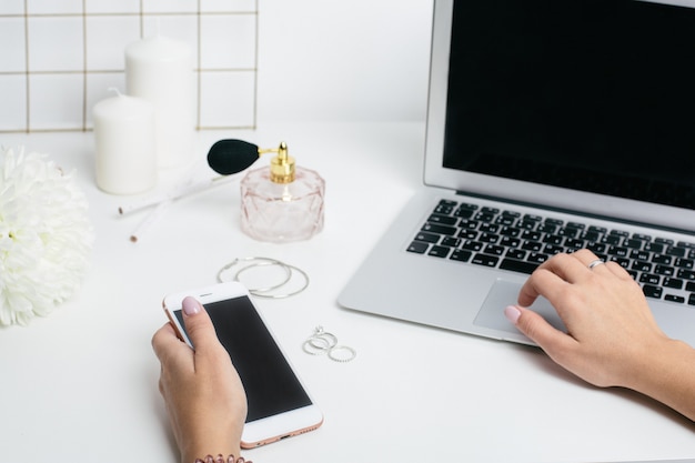 Telefone de tela em branco na mão feminina e teclado de laptop na mesa de escritório branco