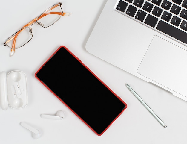 Telefone móvel vermelho, fones de ouvido e óculos perto do laptop em fundo branco