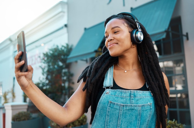 Telefone selfie e mulher negra feliz na rua da cidade com fones de ouvido ouvindo podcast de música ou rádio Tecnologia de felicidade e garota africana em uma caminhada na estrada urbana em um smartphone