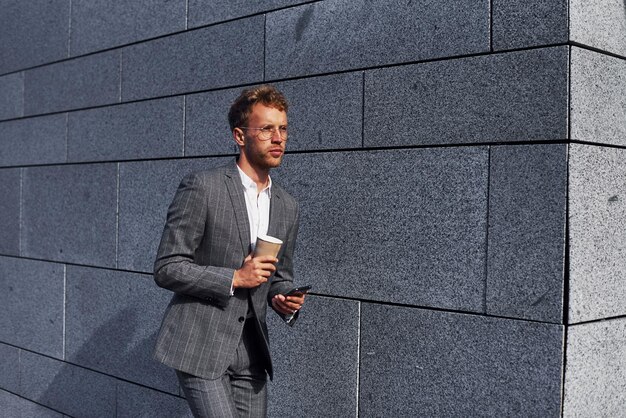 Con teléfono y bebida en las manos Joven empresario exitoso en ropa formal gris está al aire libre en la ciudad