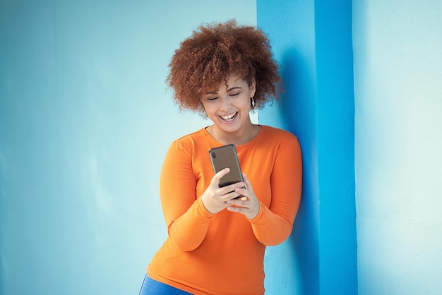 Teléfono celular de mujer negra y riendo en el móvil en la pared azul de la tecnología o red de redes sociales Niña feliz escribiendo en el teléfono inteligente de conexión leyendo notificaciones divertidas y publicaciones de memes en línea