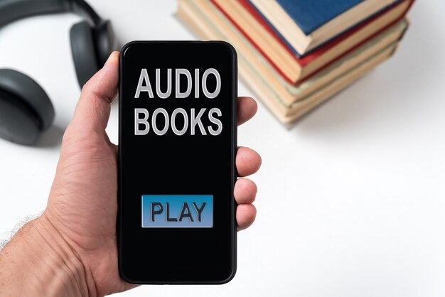 Teléfono con la inscripción audiolibros en mano en el fondo de libros y auriculares. Escuchar el audiolibro.