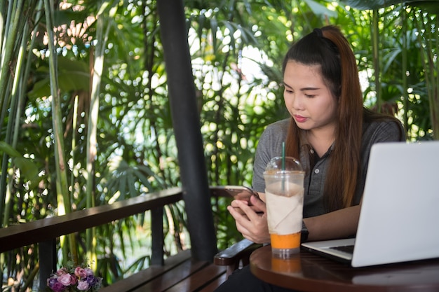 Teléfono inteligente de asimiento de la mujer joven en café del café, computadora portátil blanca y té helado de la leche