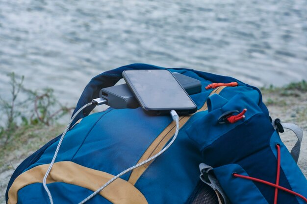 El teléfono inteligente se carga con un cargador portátil. Power Bank carga el teléfono al aire libre con una mochila para el turismo en el fondo de la naturaleza y el río.