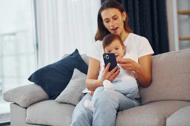Con el teléfono inteligente en la mano, la madre con su pequeña hija está en casa juntos