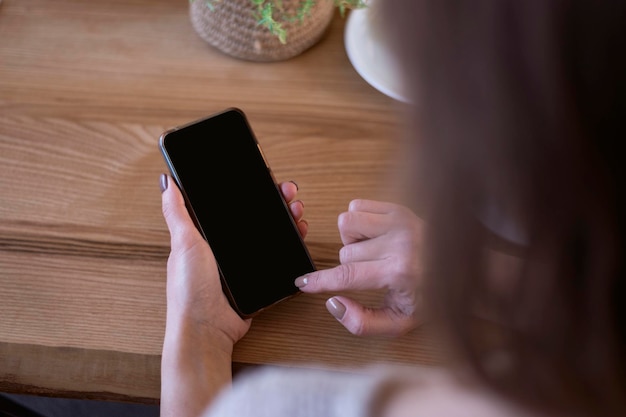 Teléfono inteligente en manos femeninas con pantalla negra en blanco Teléfono contra el fondo de la mesa de madera