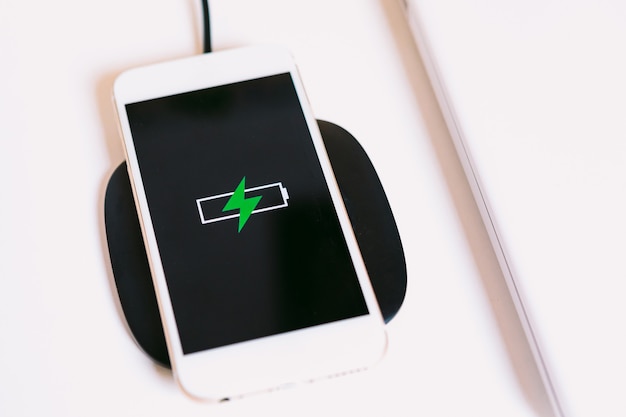 Foto teléfono inteligente móvil blanco con el logotipo de una batería con un rayo verde en la pantalla, cargando en una base de cargador sin cable junto a la computadora portátil en una mesa de trabajo blanca