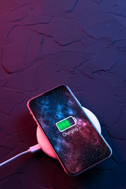 Teléfono inteligente móvil en dispositivo de carga inalámbrica sobre fondo de color rojo y azul neón oscuro. El icono de la batería y el progreso de la carga se iluminan en la pantalla. Smartphones conectados a la fuente de alimentación. Batería baja.