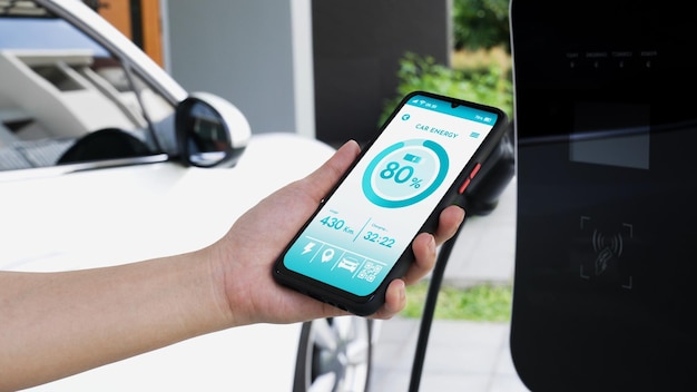 El teléfono inteligente muestra el estado de la batería en una aplicación móvil EV inteligente mientras el vehículo EV recarga electricidad mediante una estación de carga doméstica en el garaje Futuro vehículo EV innovador y sostenibilidad energética Examinar