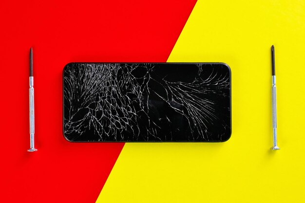 Un teléfono inteligente negro con una pantalla rota y destornilladores se encuentra sobre un fondo rojo y amarillo Vista superior