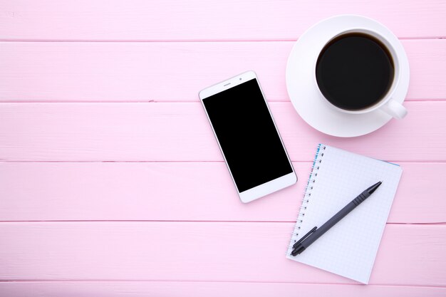 Teléfono inteligente con notebook y taza de café en madera rosa