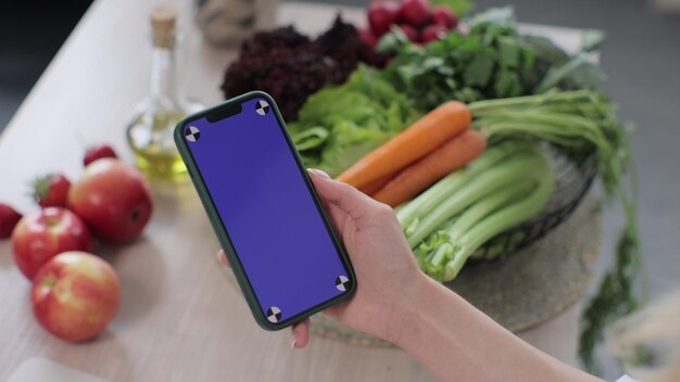 Teléfono inteligente con pantalla azul para la clave de croma en la mano de la mujer en la cocina Fondo de verduras y frutas frescas en la mesa Aplicación para cocinar Concepto de comida saludable