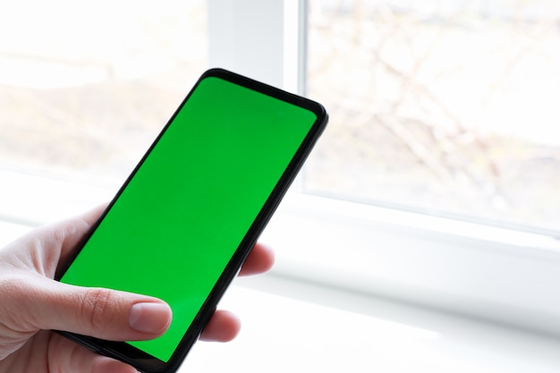 Un teléfono inteligente con una pantalla verde en la mano frente a la ventana de la habitación.