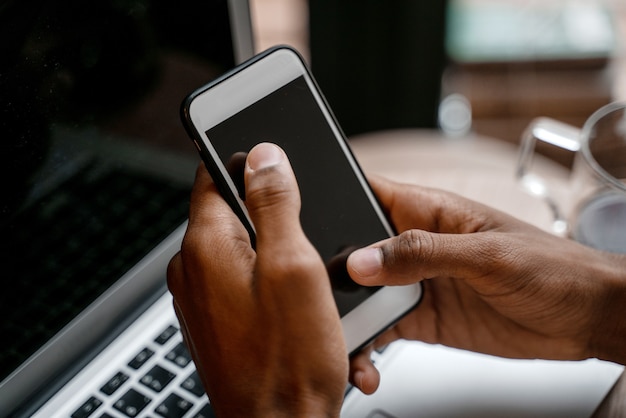 Teléfono inteligente de primer plano en manos negras con el telón de fondo de un lugar de trabajo con una computadora portátil.