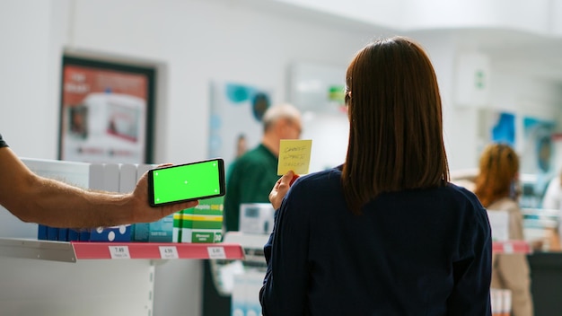 Teléfono inteligente que muestra una plantilla de pantalla verde horizontal en una tienda farmacéutica, pantalla aislada con llave cromática en el teléfono móvil. Fondo de copyspace de maqueta en blanco en el teléfono en la farmacia.