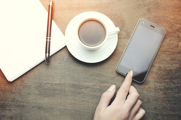 Teléfono de mano de mujer, cuaderno de mesa de madera, bolígrafo y taza de café