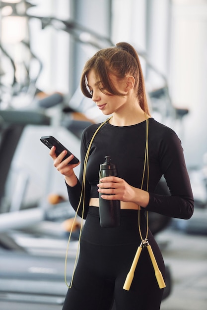 Con el teléfono en las manos y saltar la cuerda en el cuello Mujer hermosa joven en ropa deportiva tiene un día de fitness en el gimnasio